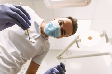 Professional dental doctor is working in office. Young man dentist in protection gloves and a mask, keeps dental instrument looking at the camera