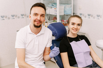 A lovely girl is sitting in the dentist's chair next to the doctor. Dentist and patient look at the camera and smile happily