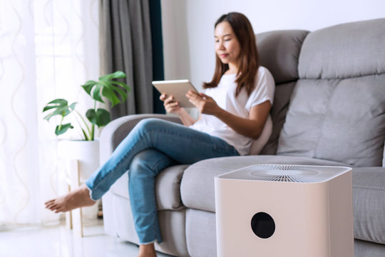 Happy Young Asian Woman Relaxed On Comfortable Sofa At Home, Wellbeing Breathing Fresh Air From Air Purifier While Dust Air Pollution Situation Outside Is Really Bad. PM 2.5, Healthy Concept.