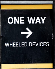 Closeup of sign that says One Way Wheeled Devices with an arrow pointing to the right.