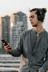 Man sitting on the rooftop and listening music application in wireless black headphones and checking his smartphone