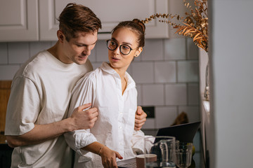happy caucasian married couple in the kitchen, at morning. woman work on laptop, she is at freelance work, they are in domestic clothes. relationships, home, love, support, work