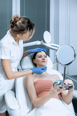 Young woman at beauty clinic cosmetology service sitting on medical chair while female doctor examining skin with mirror