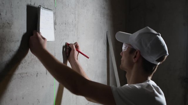 the master of decorating makes notes on the wall using a pencil and notes in his notebook. Young builder is finishing in a client s apartment