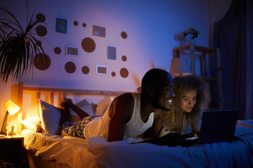 Full length portrait of young mixed-race couple using laptop while lying in bed at night, copy space