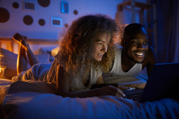 Portrait of young mixed-race couple using laptop and smiling while lying in bed at night, copy space