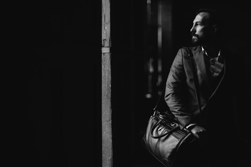 A man with a beard in a suit and with a travel bag is in a dark room. Black-and-white photografy