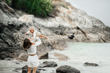 A young slender mother throws her baby up in her arms with joy against the backdrop of a rocky beach and waters. motherhood and Childhood.