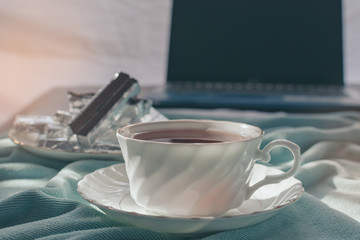 A cup of tea on the bed and light blue blanket with sunrise, Morning teatime with chocolate, notebook computer,smartphone, Normal practice by working at home during covid-19 outbreak around the world.
