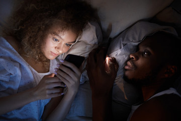Above view at young mixed-race couple using smartphones while lying in bed at night, focus on smiling curly-haired woman
