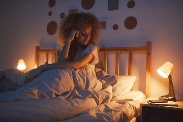 Portrait of curly-haired young woman sitting in comfortable bed at night and looking at smartphone while waiting for call or text message, copy space