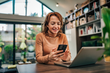 Fototapeta premium Happy woman using smart phone and laptop at home, portrait.