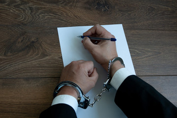 A man in handcuffs sits at a table in front of a blank sheet of paper and holds a fountain pen....