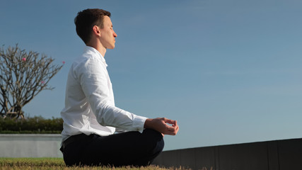 young businessman in white shirt meditates sitting in lotus pose on hotel terrace at sunlight against blue sky side view