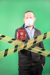 A man in a protective medical mask on a green background holds a passport in his hand. Yellow forbidden tape in front of a standing person. Closing borders due to the epidemic of coronavirus.