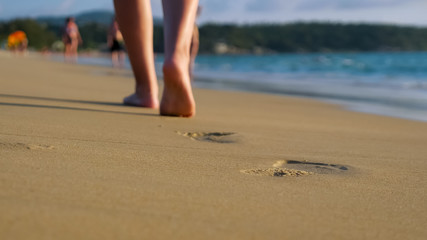 Fototapeta na wymiar barefoot lady walks on empty ocean beach wet sand leaving footprints under blue sky on sunny day close low angle backside shot