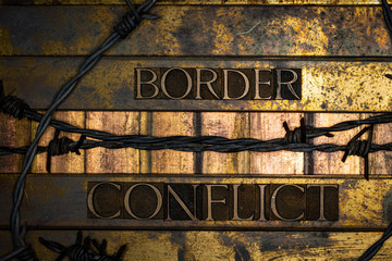 Photo of real authentic typeset letters forming Border Conflict text with barb wire on vintage textured silver grunge copper and gold background
