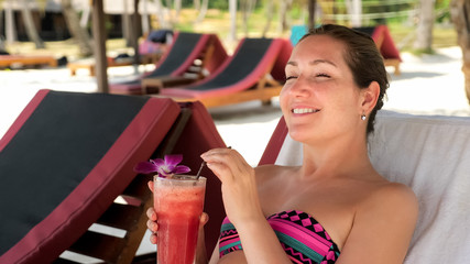 smiling girl in bikini lies on sunbed and drinks delicious cocktail under beach shed enjoying vacation in tropics close view
