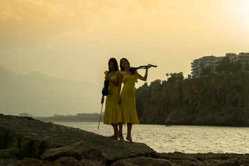 Two beautiful girls violinists in yellow concert dresses are playing electric violins on sunset on...