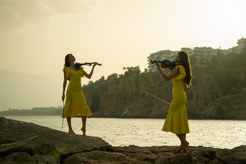 Two beautiful twin sisters violinists in yellow concert dresses are playing electric violins on...