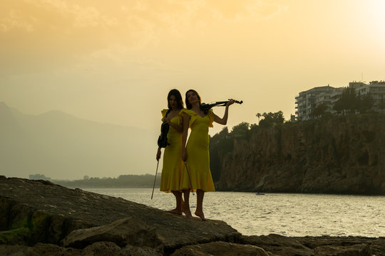 Two beautiful girls violinists in yellow concert dresses are playing electric violins on sunset on rocks by the Mediterranean sea in old town of Antalya Kaleici Turkey.