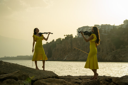 Two beautiful twin sisters violinists in yellow concert dresses are playing electric violins on sunset on rocks by the Mediterranean sea in old town of Antalya Kaleici Turkey.