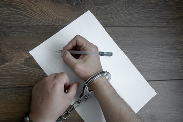 Hands of the criminal in handcuffs write with a pen on paper. Sincere recognition, request,...
