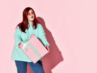 Fototapeta premium Young red haired overweight woman in jeans, blue hoodie and sunglasses standing with gift box and expressing surprise