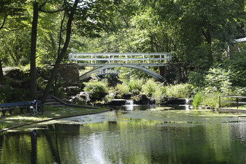 silent pond in the shadow of massive greens trees and a pedestrian white bridge over it. Little low waterfalls near a pond in lush green summer environment