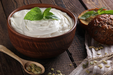 Homemade sour cream in a deep clay bowl with herbs, basil, bread and rye ears on an old wooden background. Rustic. Spoon with pumpkin seeds. Background image, copy space