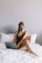 Young female in silk sleepwear and with mud mask on face sitting on soft bed and browsing laptop during skin care routine at home