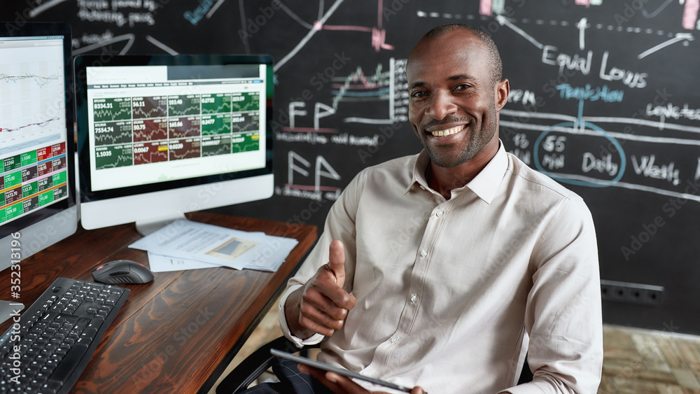 Poster Money decide your level. Cheerful african american male trader sitting by desk, smiling at camera, showing OK sign and studying analytical reports using tablet pc in the office