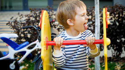 Funny portrait of little boy riding on chidlren playground and showing tongue