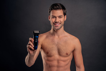 Handsome young bearded man isolated. Portrait of topless muscular man is standing on gray background with electric Shaver in hand. Men care concept.
