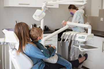 The boy on his mother's lap sitting on the dental chair, Due the Covid-19 pandemic they have face...