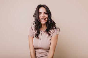 Portrait of cute dark-haired girl with snow-white smile. Brown-eyed lady in beige top looking at camera