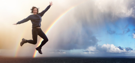 Fototapeta na wymiar Happy and Excited Girl Jumping with Joy. Ocean Background with Rainbow and Rain Clouds.
