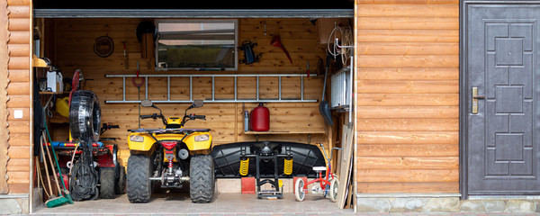 Facade front view open door ATV quad bike motorcycle parking messy garage building with wooden siding at home driveway backyard and lawn path. House warehouse for tools and equipment . Wide panoramic - Powered by Adobe