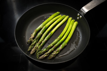 Fresh green asparagus is roasted in a black pan on the stove, copy space