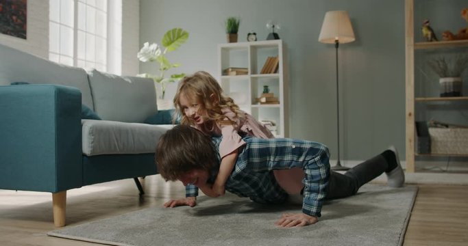Funny joyful little caucasian girl having a piggy back ride on her father while he is doing push ups, spending time at home together - real people, family home 4k footage