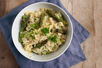 Risotto with green asparagus, parmesan and parsley garnish in a white bowl on a napkin and a rustic wooden table, copy space, high angle view from above