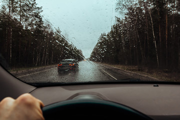 View through windshield of car on drops on glass, wet road and rainy weather. Highway car traffic in bad weather, downpour. Dangerous overtaking. Dashboard inside and driver hand with steering wheel
