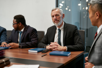 Busy working day. Portrait of bearded mature man in formal wear looking at camera while sitting at the office table with his co workers in the modern office