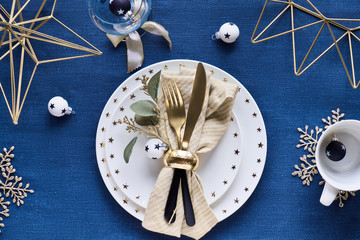 Christmas table setup with white plate, golden utensils and gilded geometric metal decor and gilded snowflakes. Flat lay on dark blue linen textile table cloth background.