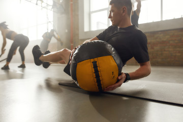 Sportive man using exercise ball while having workout at industrial gym. Group training, teamwork...