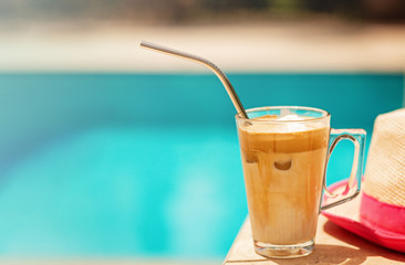 Ice coffee Fredo against blue clear water of the swimming pool with straw hat