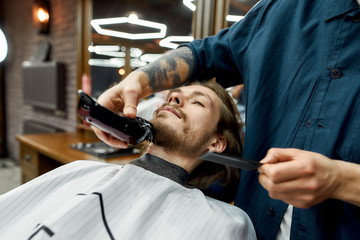 Beauty day for men. Young guy sitting in armchair in the barbershop while barber with tattoo on arm trimming his beard. Working with electric clipper machine