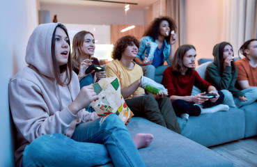 Having fun at home. Young multicultural people playing video games, eating snacks and relaxing...