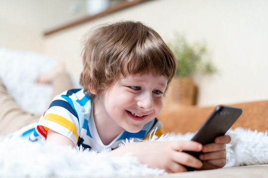 Happy little child boy playing online game, watching video on cellphone, lying on couch entertaining in living room. Smiling small kid using funny mobile apps, enjoying free leisure time at home