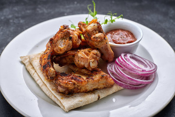 Grilled chicken wings with barbecue sauce, pita bread, microgreen and onion rings, on a white plate, against a dark background